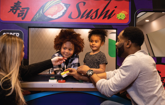 Two children in a model food truck serve toy sushi to two adults.