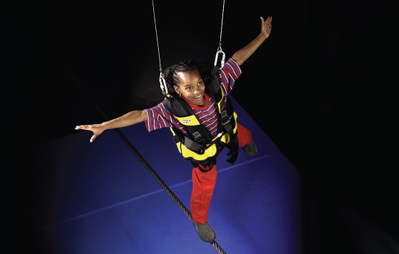 A child wearing a full safety harness, walks a tightrope.