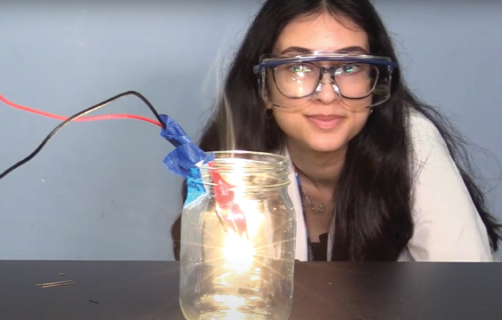 An educator wearing safety goggles monitors a flame inside a jar with crocodile clips attached to it.