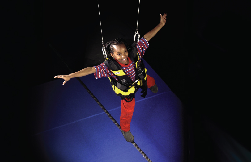 A child wearing a full safety harness, walks a tightrope.