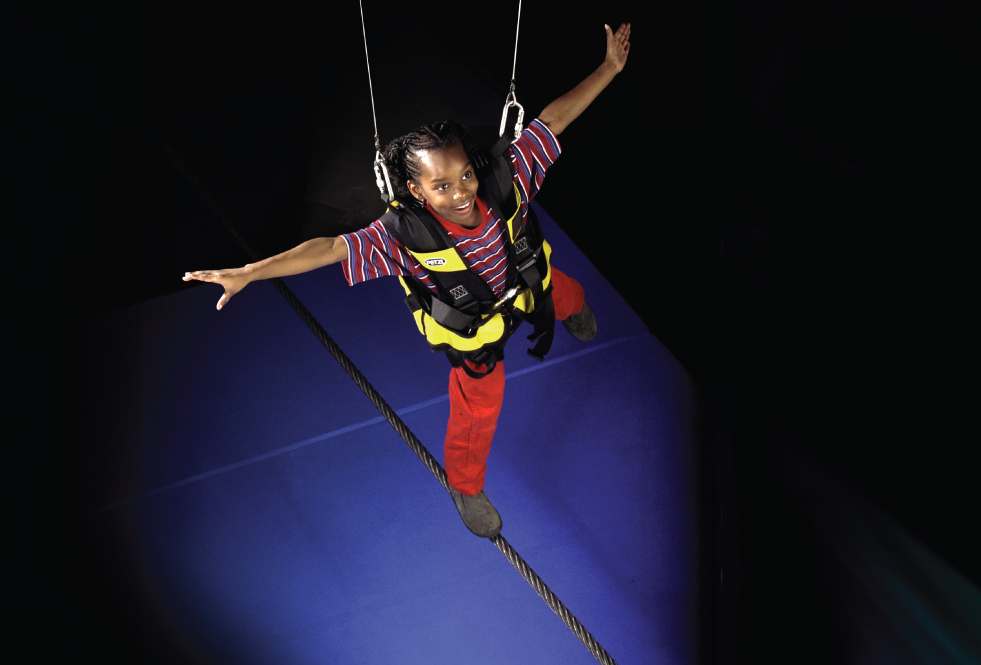 A child wearing a full safety harness, walks a tightrope.